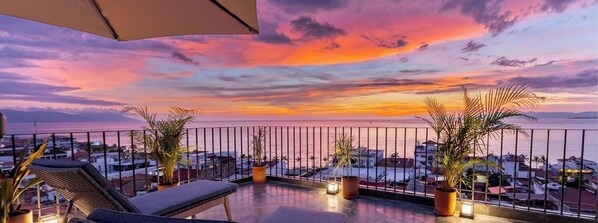 A balcony with a view of the sunset over the sea, potted plants, and a lounge chair.