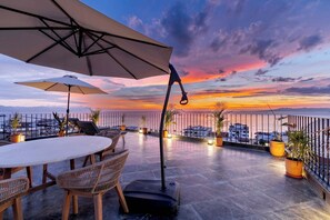 A rooftop terrace with a round table and chairs, potted plants, and umbrellas.