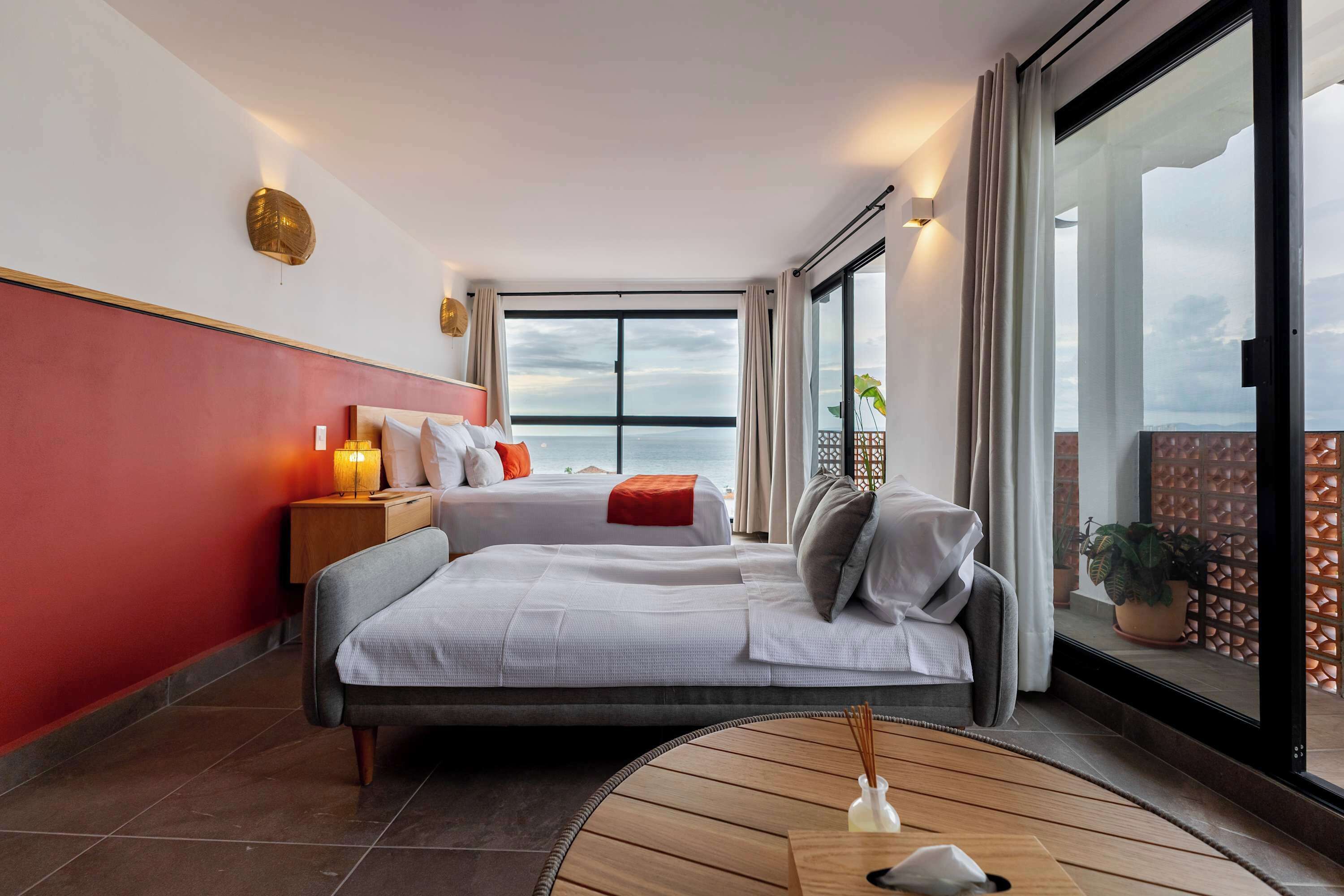 Una habitación de hotel moderna con una cama grande, una mesita de noche con lámpara, vistas al mar a través de amplios ventanales y un balcón con plantas en macetas.