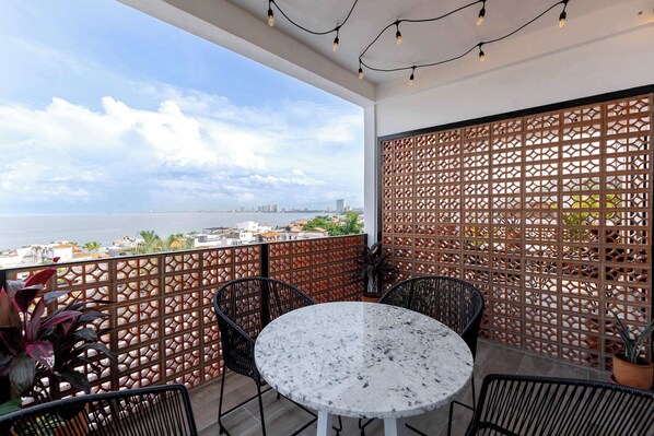 A balcony with a white table and chairs, a wooden lattice partition, and a view of the sea and cityscape.