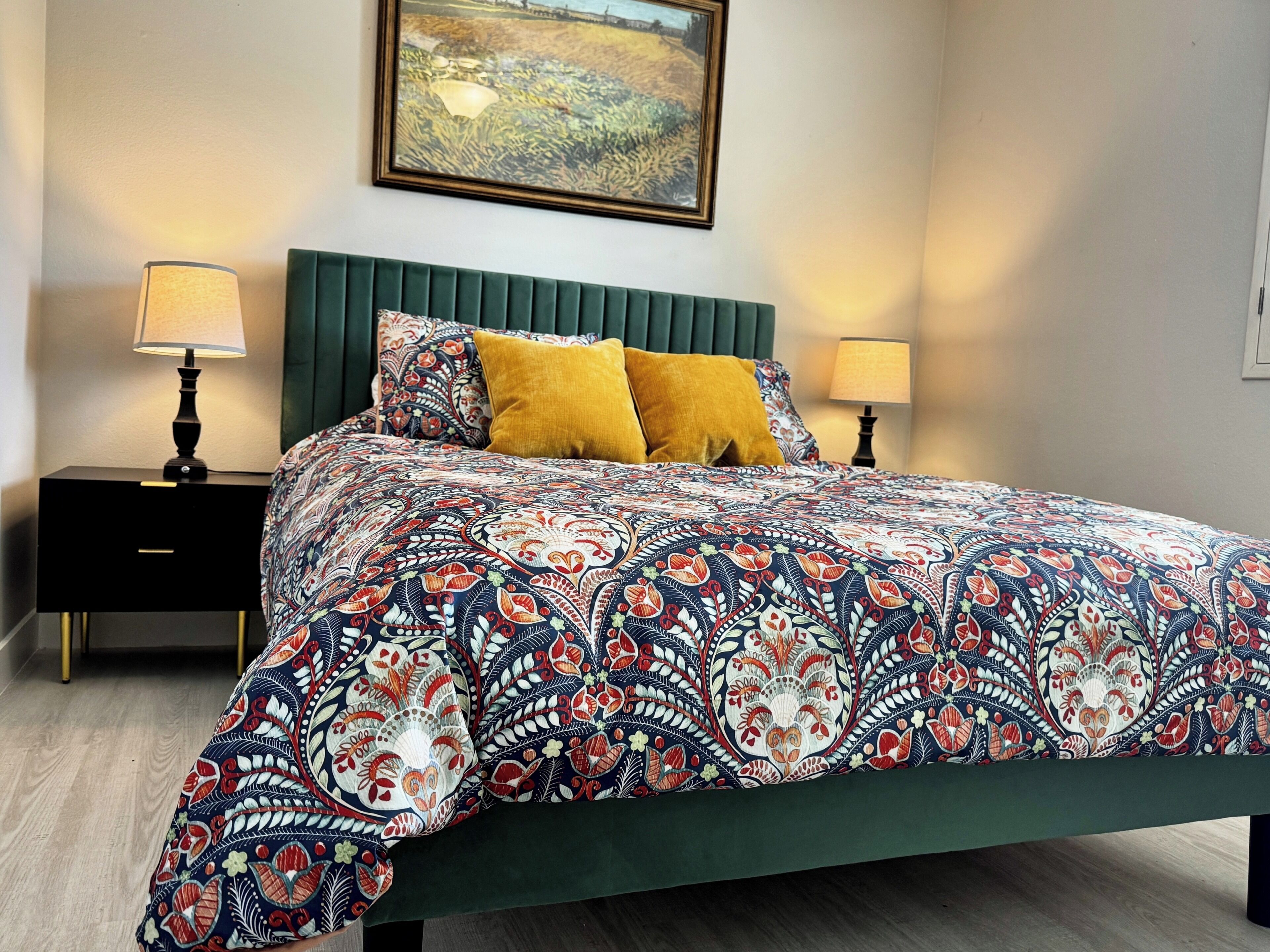 A bedroom with a bed, green headboard, two nightstands with lamps, a painting on the wall, and a patterned bedspread.