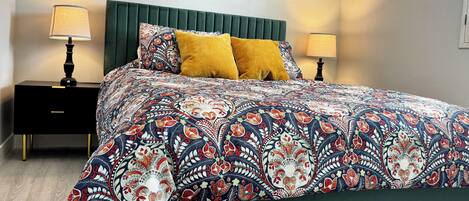 A bedroom with a bed, green headboard, two nightstands with lamps, a painting on the wall, and a patterned bedspread.