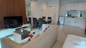 Un salon moderne avec un canapé, une table basse et un téléviseur. On y trouve également un coin repas avec des chaises et une table. La cuisine est visible, avec des meubles blancs et un plan de travail.