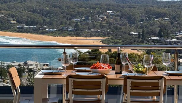 A dining table with chairs and wine glasses, overlooking a beach and forested hills.