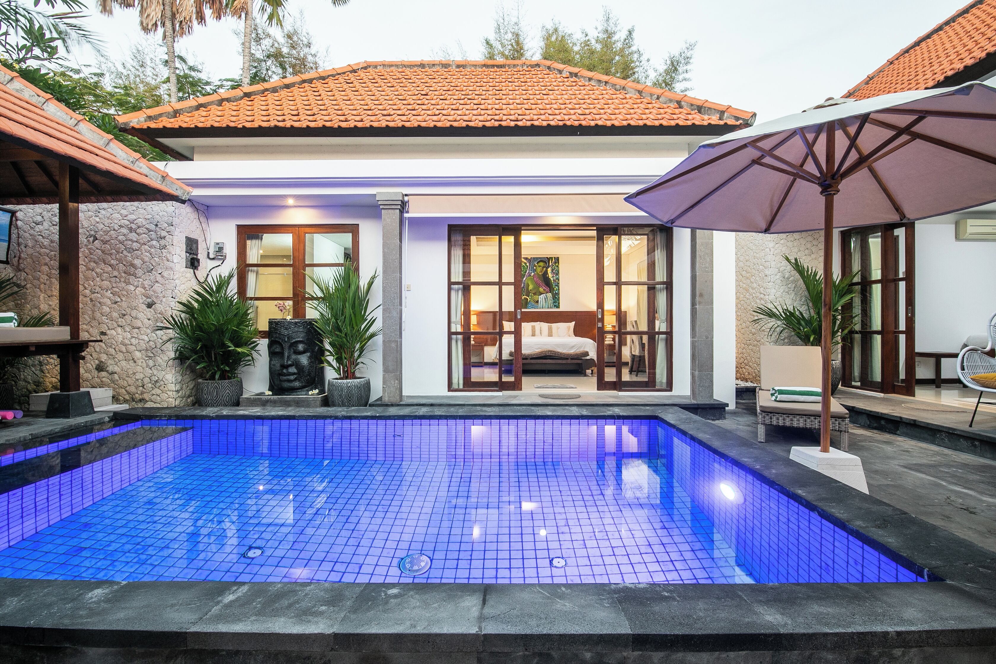 A swimming pool with a tiled blue floor, a covered patio area with a chair and umbrella, and a building with a tiled roof and large windows.