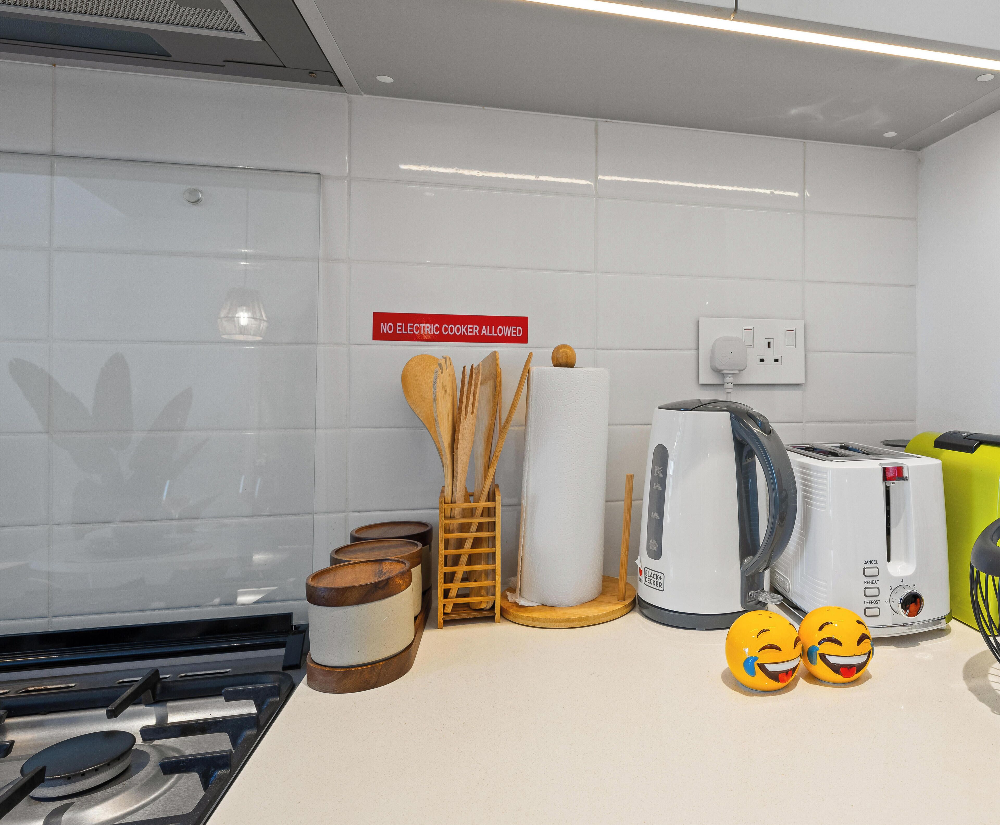 A modern kitchen with white tiles, a gas stove, a kettle, a toaster, and a coffee maker. There is a sign prohibiting electric cookers.