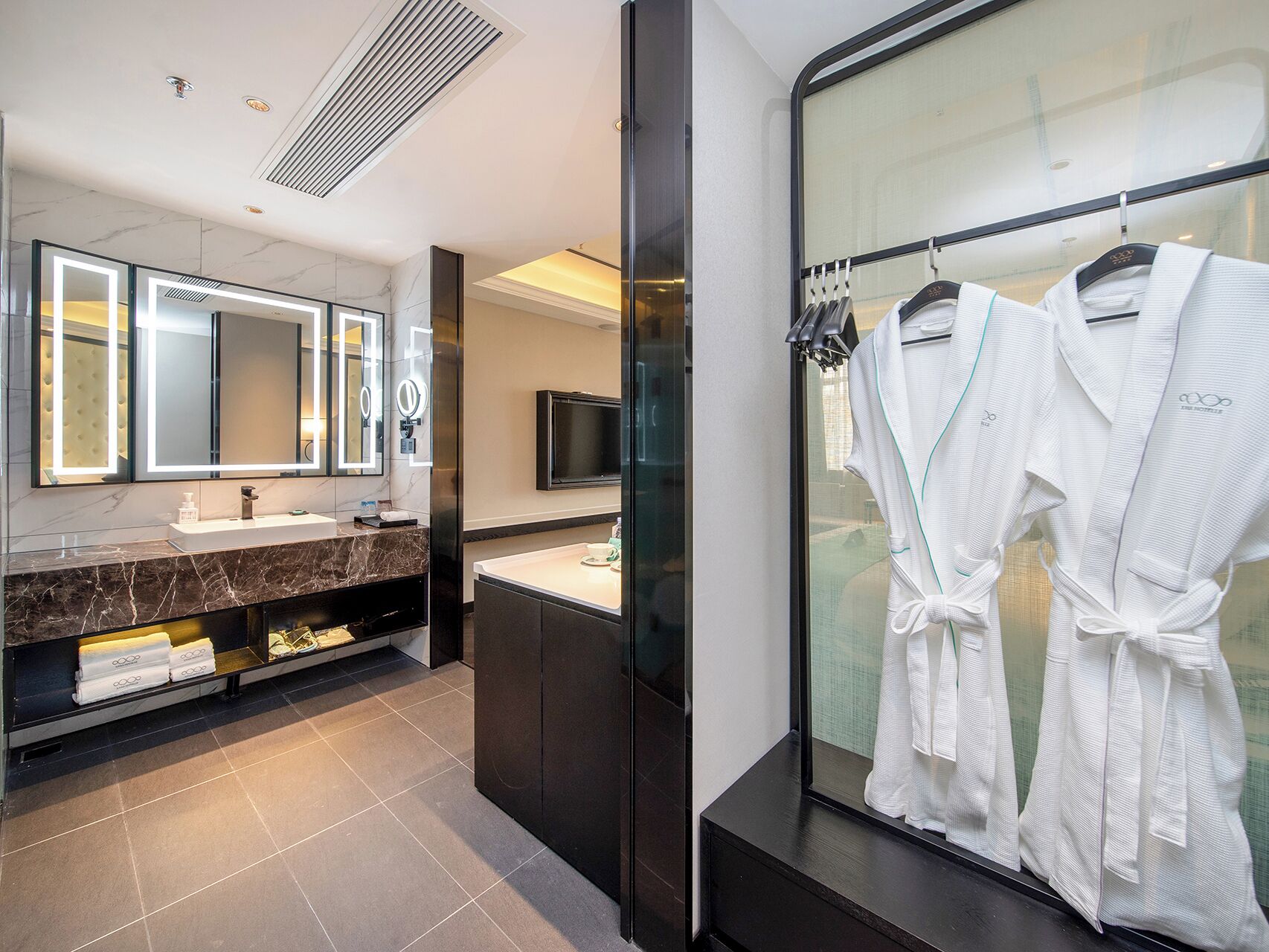 A modern bathroom with a marble vanity, a large mirror with integrated lighting, a television, and a glass shower enclosure with white robes hanging on a rack.