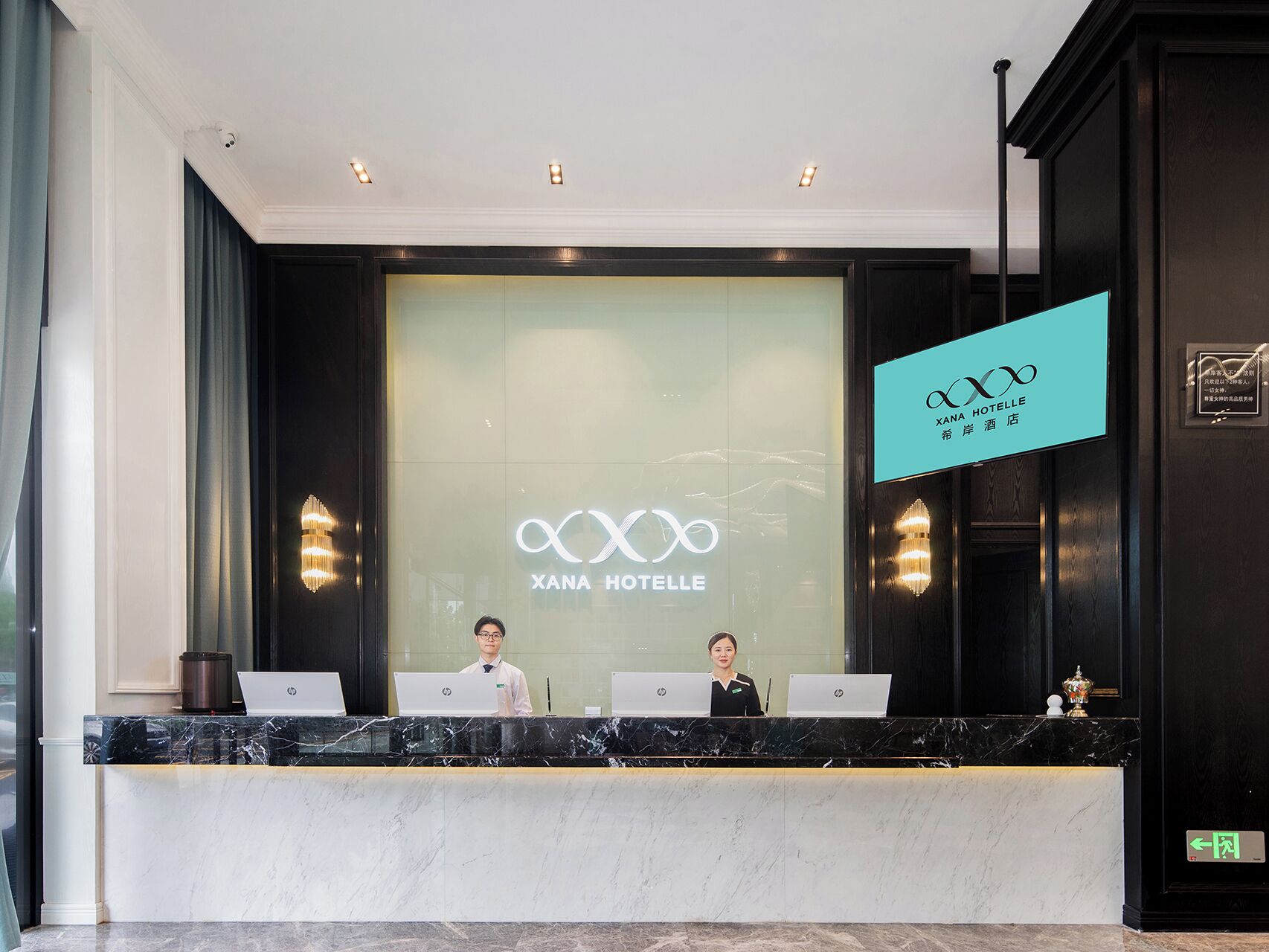 Στο XANA HOTEL υπάρχει ένας πάγκος υποδοχής, πίσω από τον οποίο βρίσκονται τρία μέλη του προσωπικού, καθένα από τα οποία έχει έναν φορητό υπολογιστή.