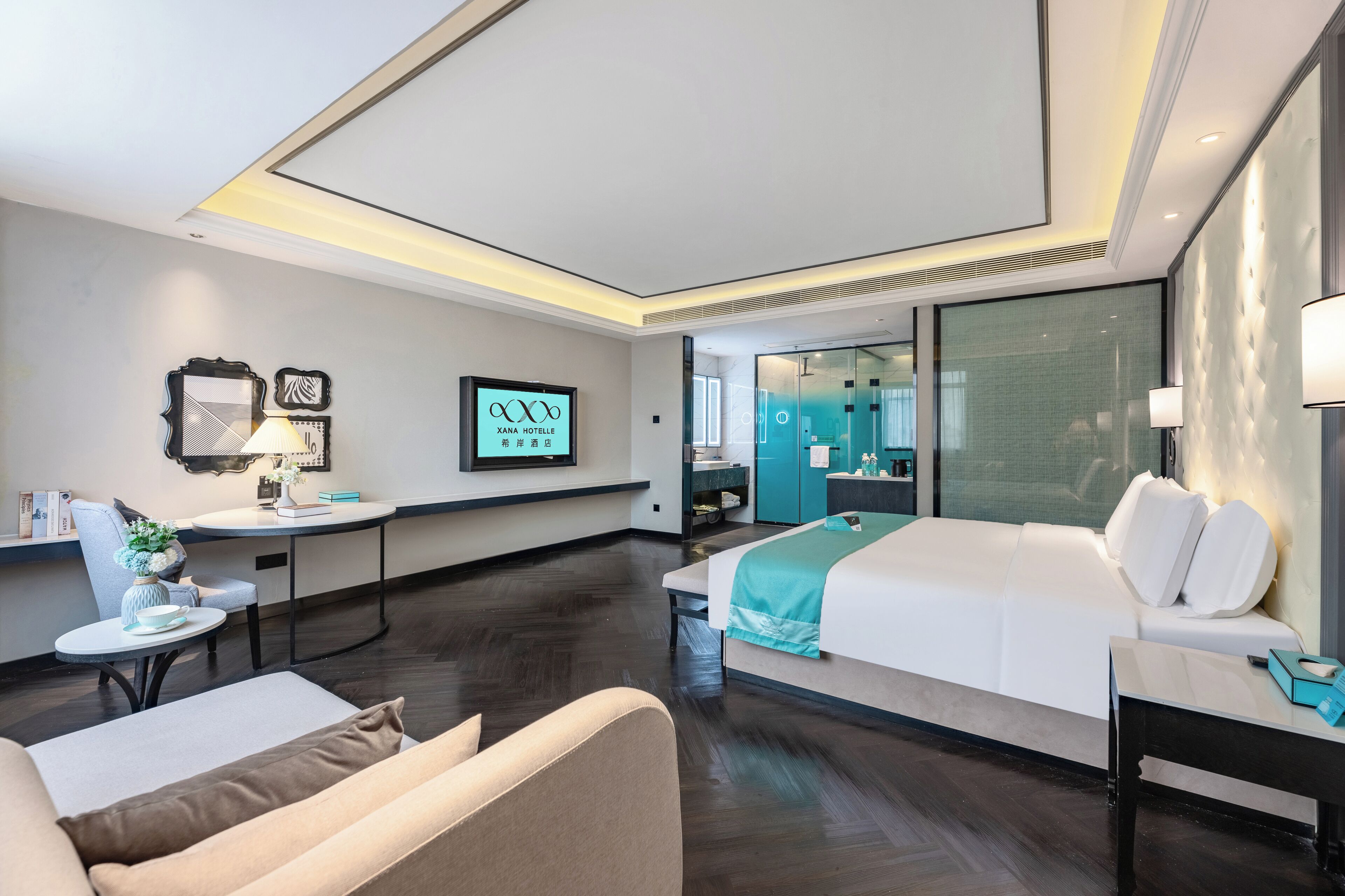 A hotel room with a bed, a desk, a chair, a small table with a vase, and a TV screen displaying the hotel name.