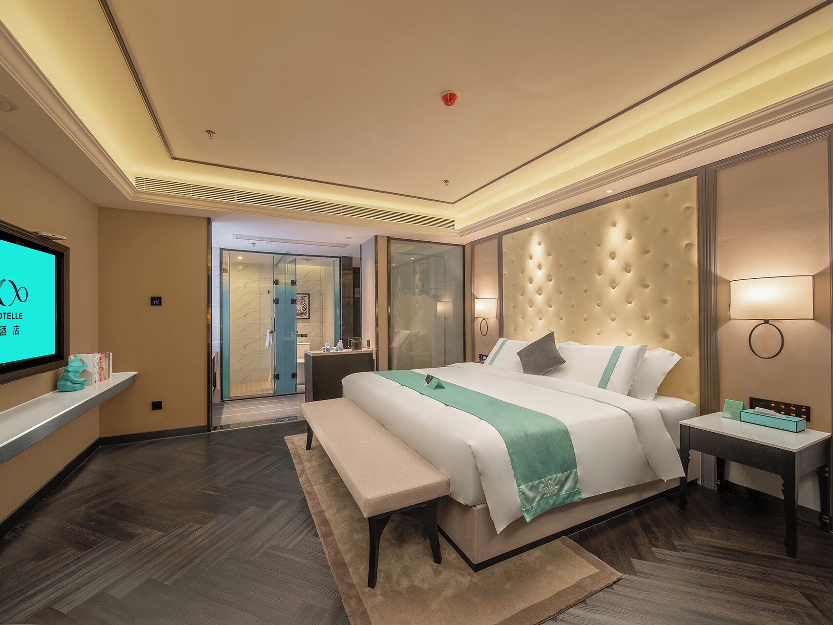 A modern hotel room with a large bed, a bedside table, a TV, and a bathroom visible through a glass door.