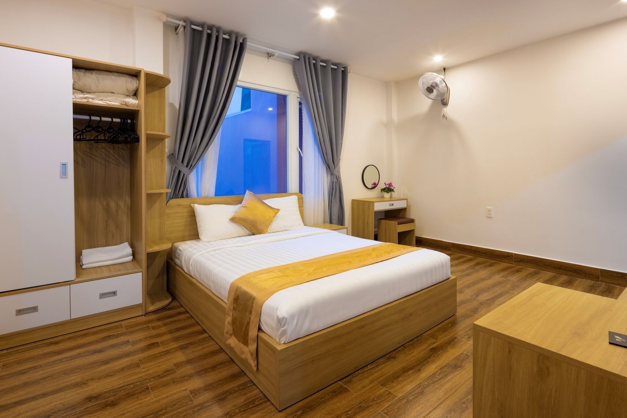 A modern hotel room with a wooden bed, a wardrobe, a desk, and a window with curtains.