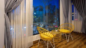 A room with a wooden floor, two yellow wicker chairs, a small round table with a vase, and a view of a night scene outside through large windows with sheer curtains. - Romance Hotel (Lam Vien - Da Lat)