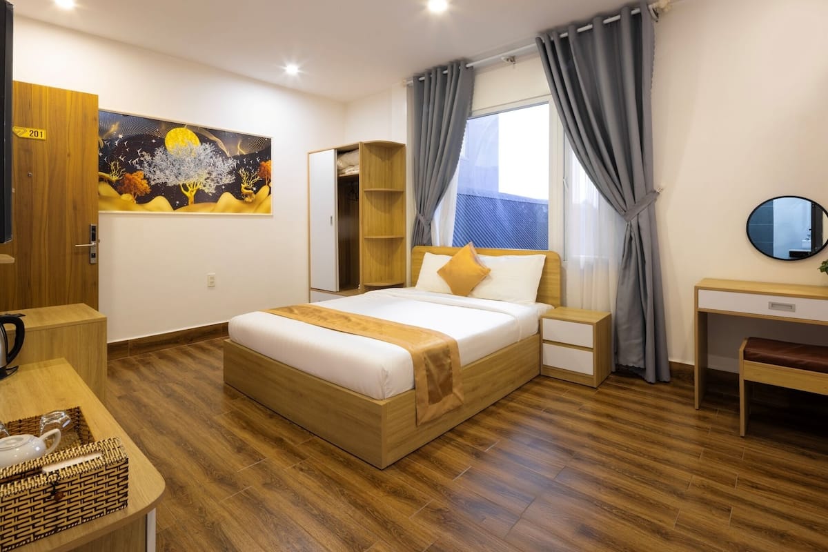 A hotel room with a wooden floor, a bed with white bedding, a desk, a chair, a wardrobe, a painting on the wall, and a window with curtains.