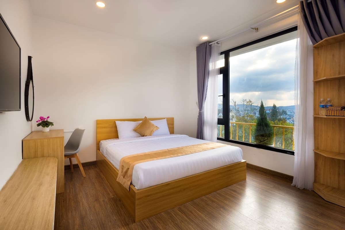 A modern hotel room with a wooden bed, a desk, a chair, and a large window offering a city view.