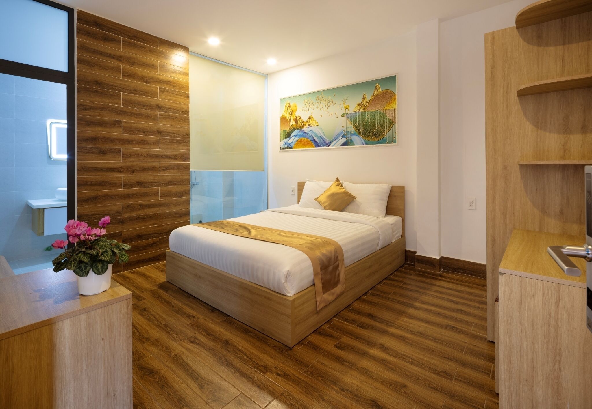 A modern bedroom with a wooden floor, a bed with white bedding, a painting on the wall, and a desk with a plant.