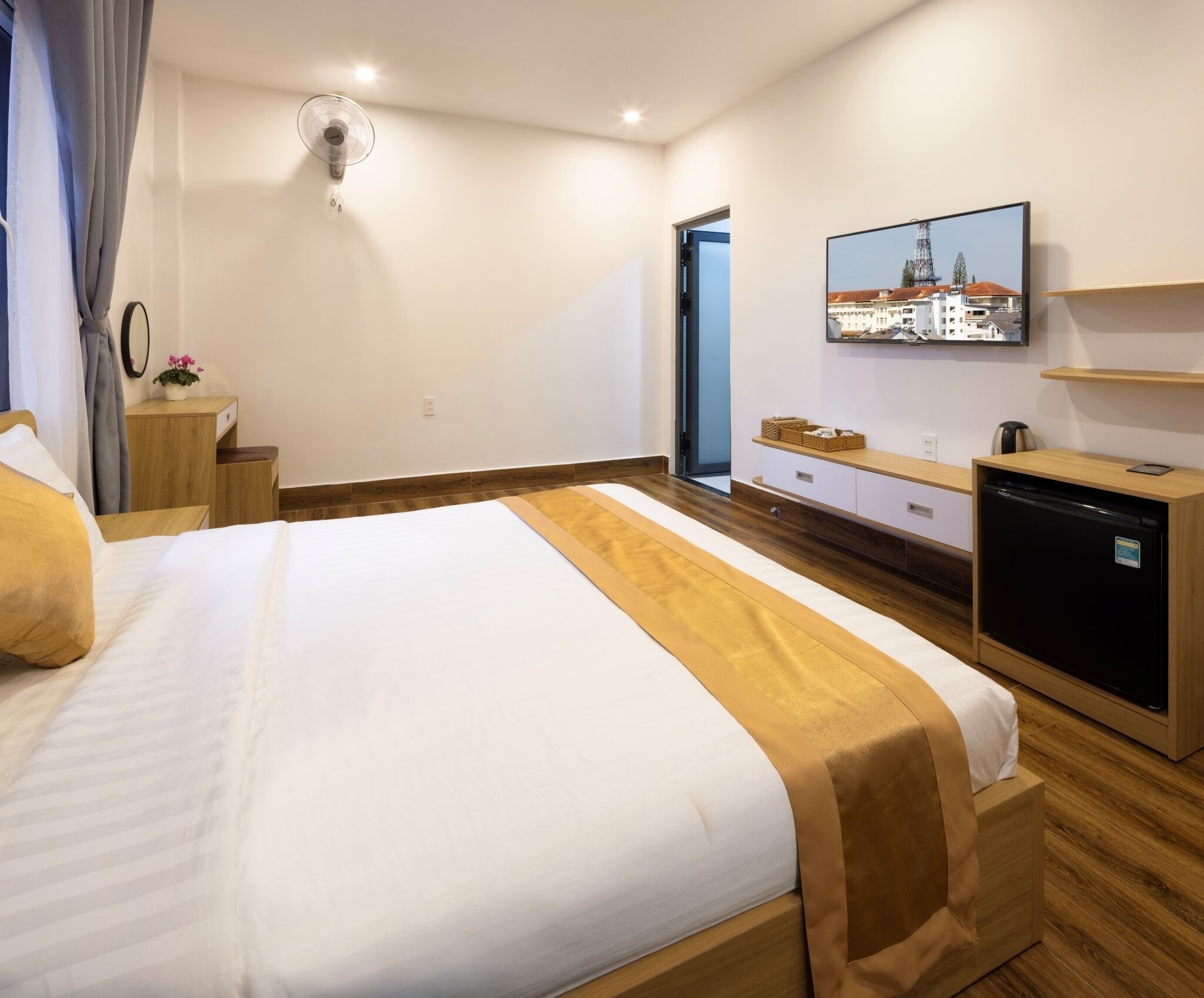 A hotel room with a bed, a TV mounted on the wall, a wooden dresser, and a small table with a plant.