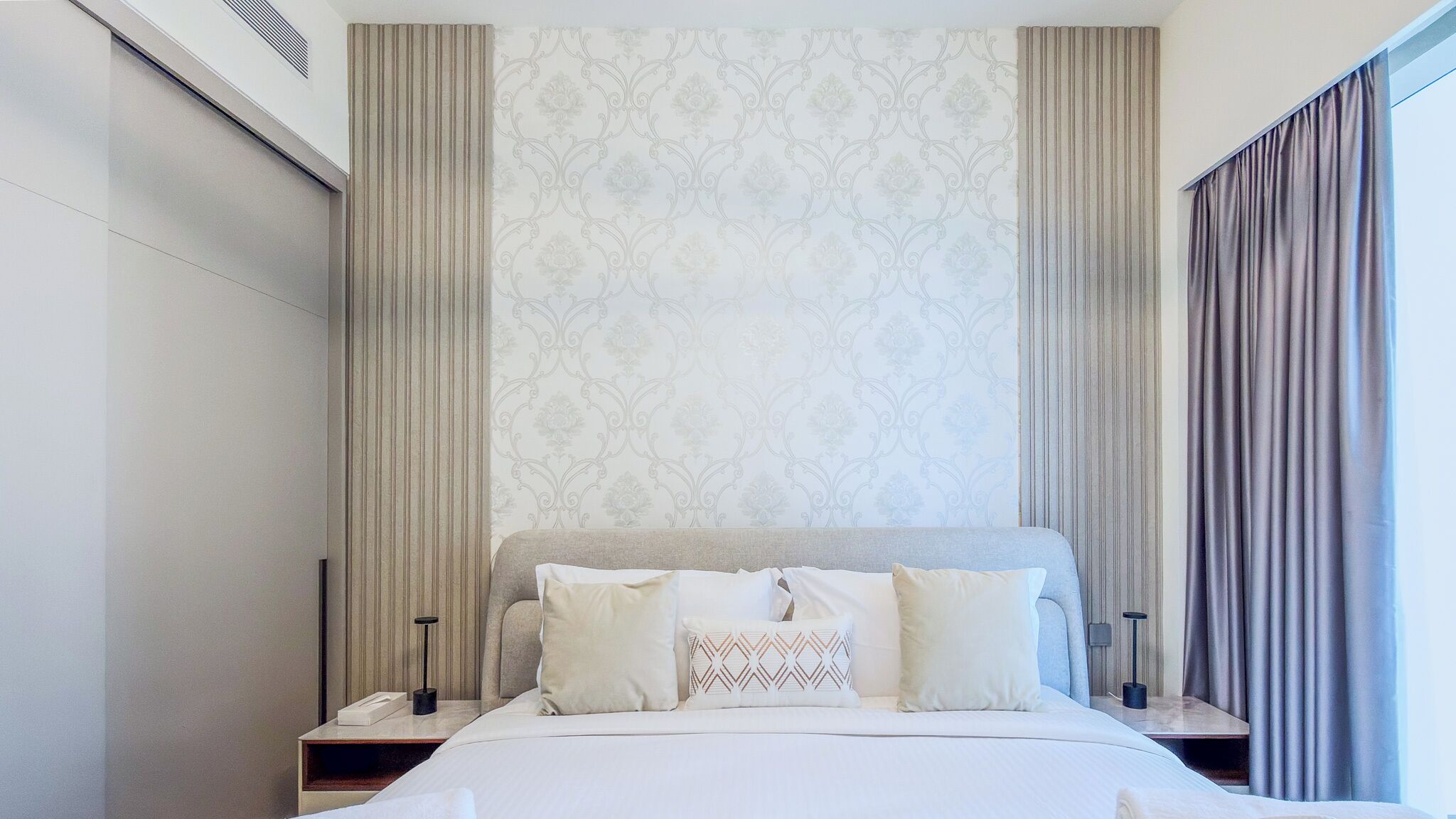 Une chambre d’hôtel moderne avec un grand lit, deux tables de chevet, une petite table d’appoint, un papier peint à motifs et une fenêtre avec des rideaux.