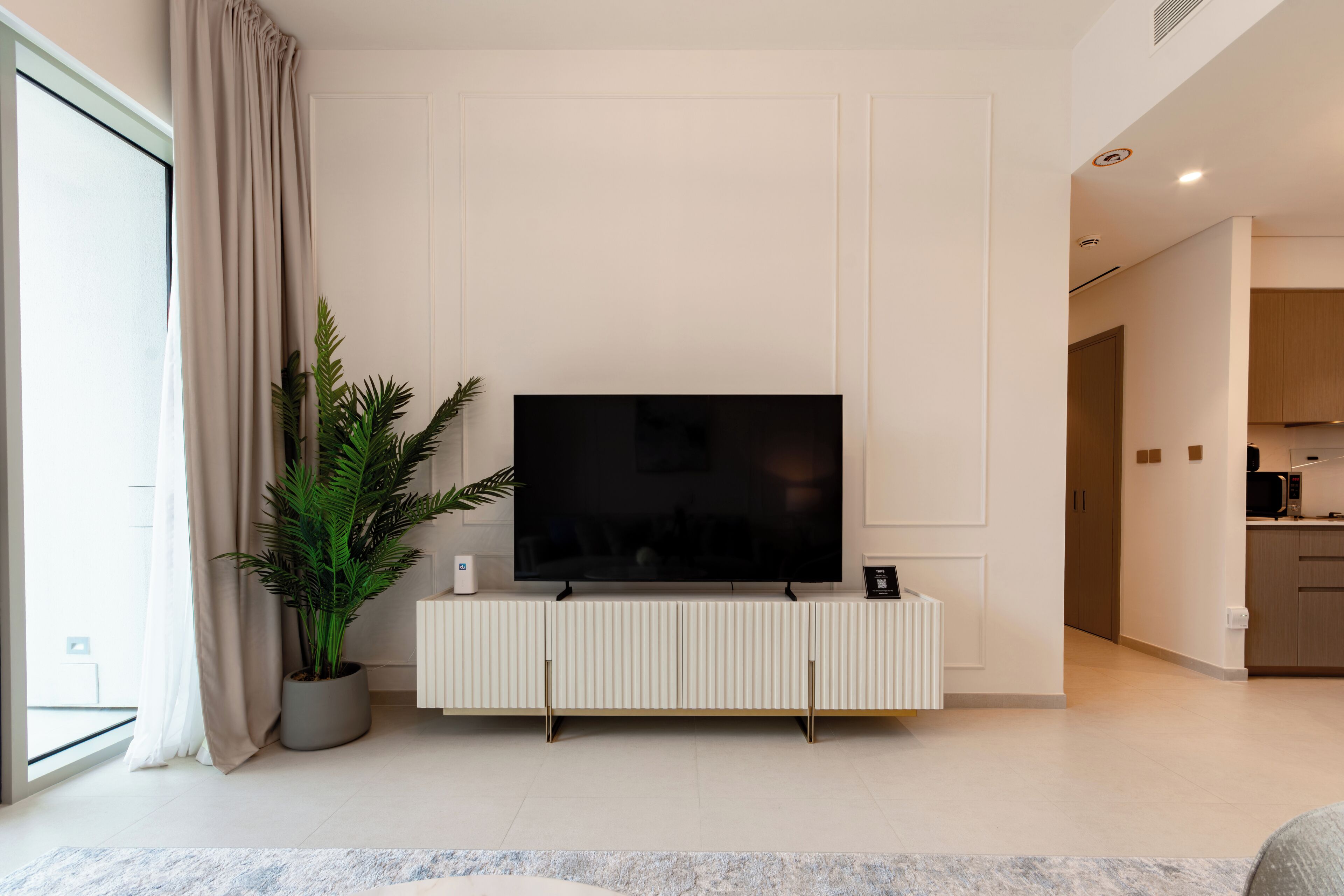 A modern living room with a large flat-screen TV on a white console, a potted plant, and a view into the adjacent kitchen.