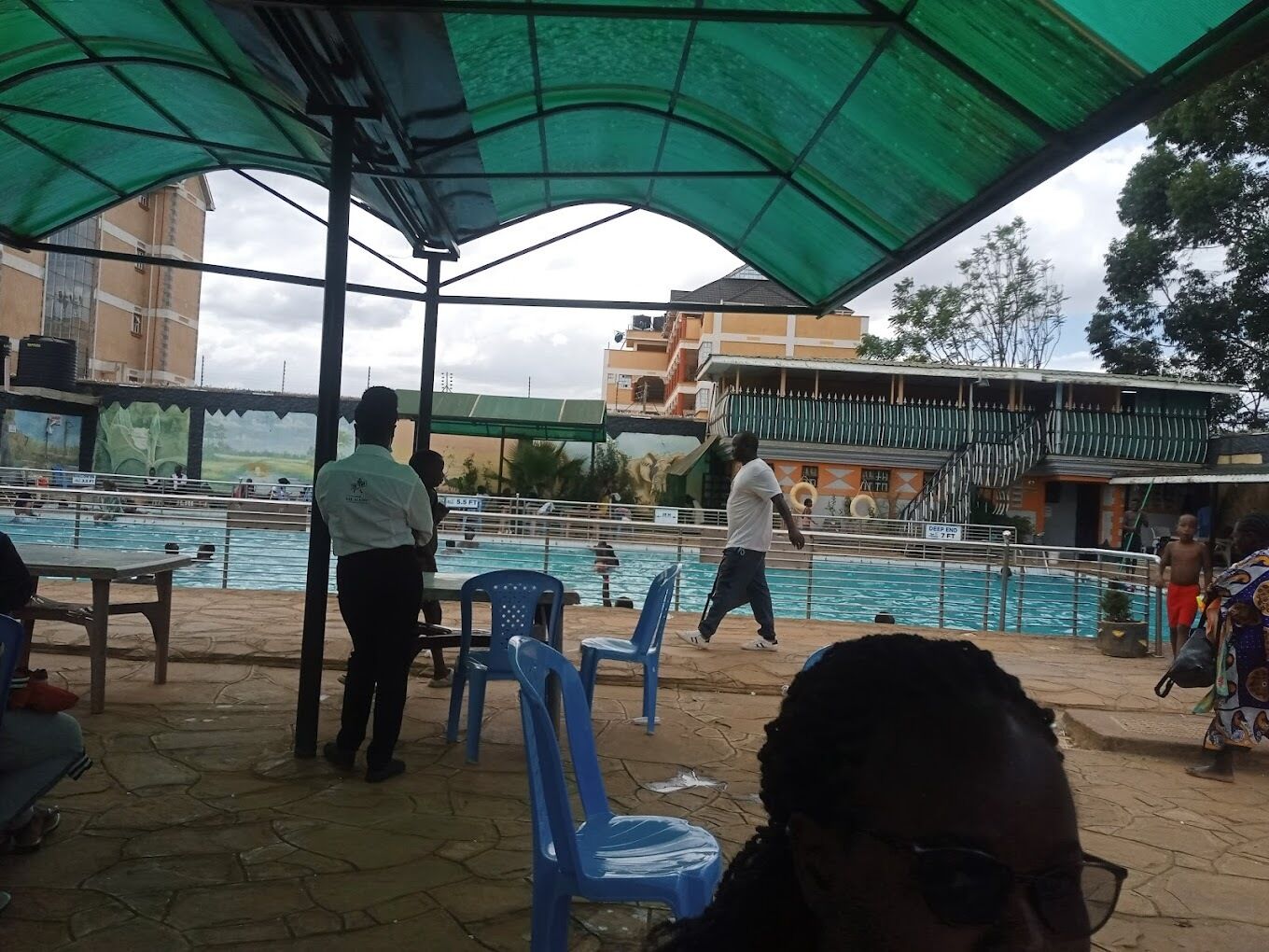 Zona de piscina exterior con área de estar techada, socorrista de servicio y un edificio con balcón al fondo.