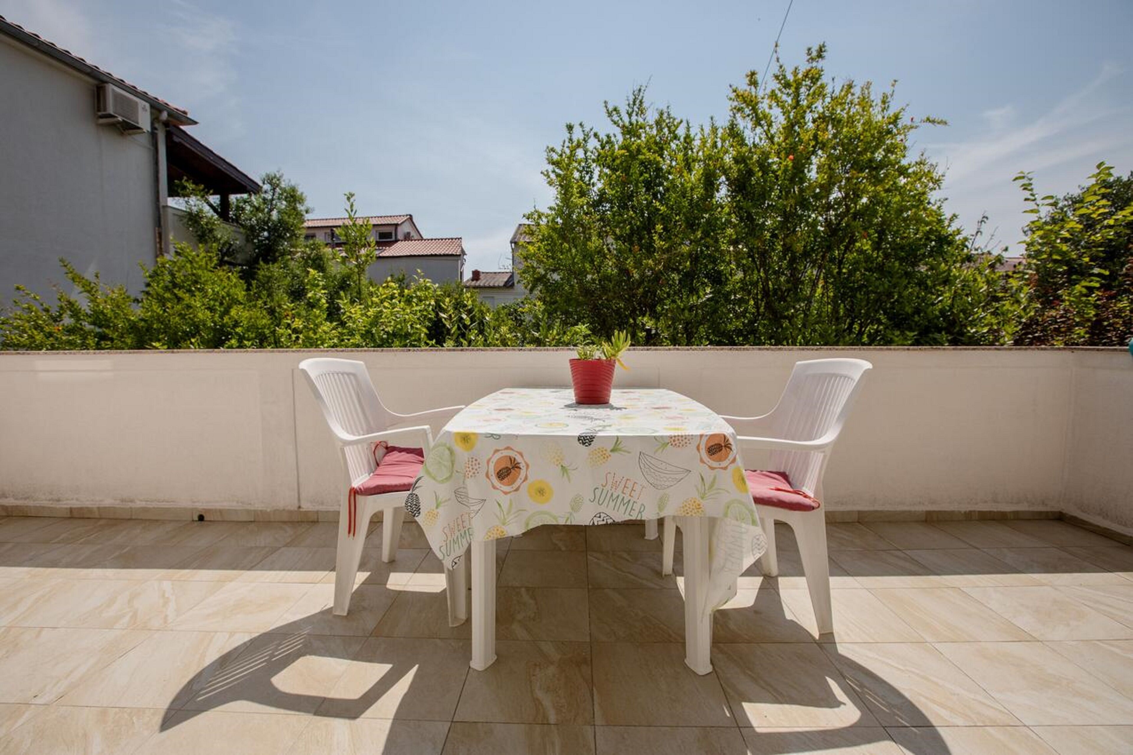 Une petite terrasse extérieure avec une table et deux chaises, un pot de fleurs et une vue sur un quartier résidentiel avec des arbres et des immeubles.