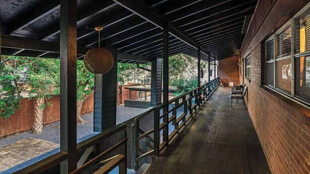 Un espacio al aire libre techado con vigas de madera y barandilla de metal, con un muro de ladrillo y ventanas en un lateral.
