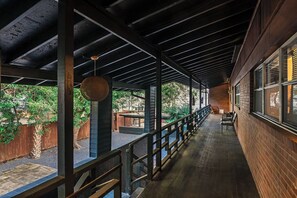 Un espacio al aire libre techado con vigas de madera y barandilla de metal, con un muro de ladrillo y ventanas en un lateral.
