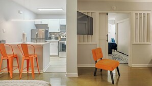 Una cocina moderna con armarios blancos, un refrigerador de acero inoxidable y un horno empotrado. Hay dos sillas naranjas en primer plano y una silla negra al fondo.