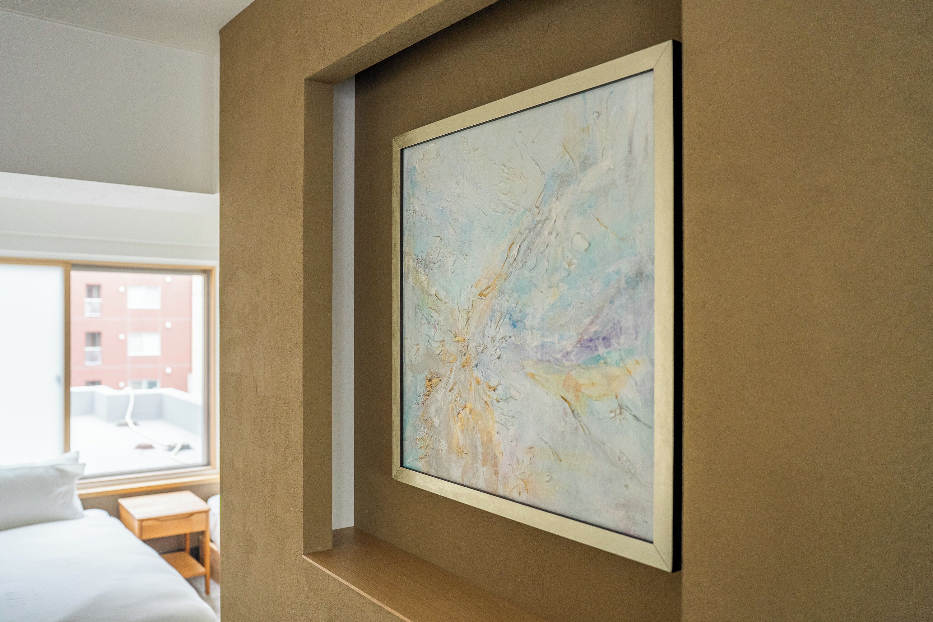 Habitación de hotel con un cuadro abstracto grande en la pared, una cama, una mesita de madera y una ventana con vistas a edificios.