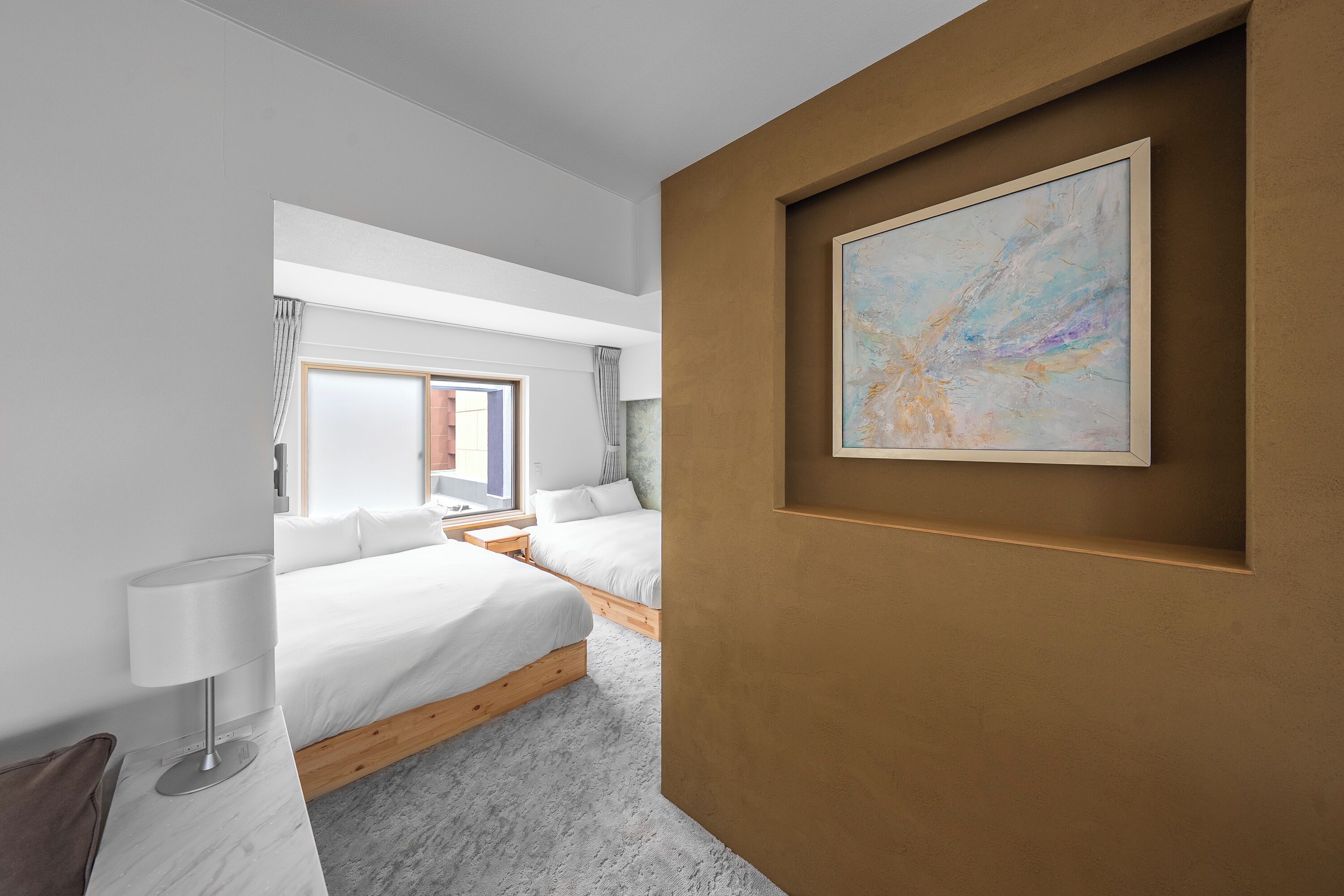 A modern bedroom with two beds, a large window, and a painting on the wall.