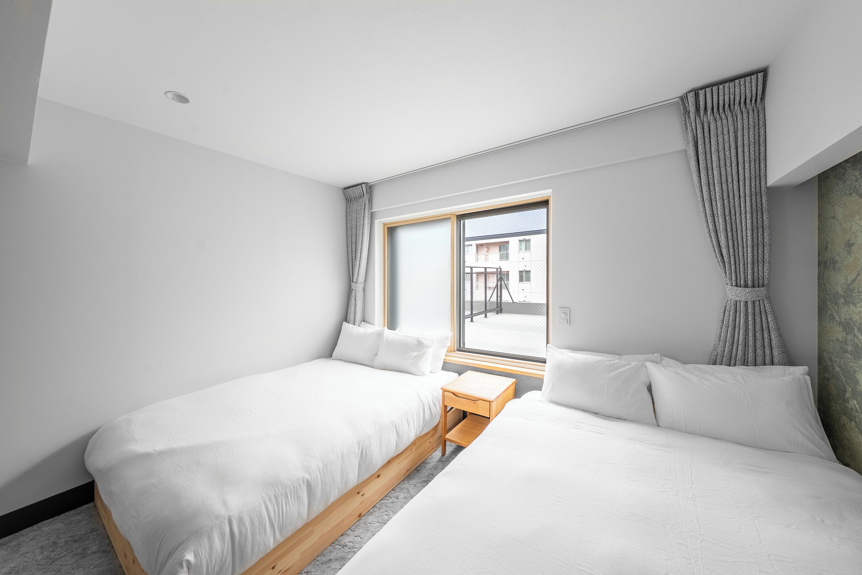 A modern hotel room with two single beds, a small wooden table, and a large window offering a view of the outside.