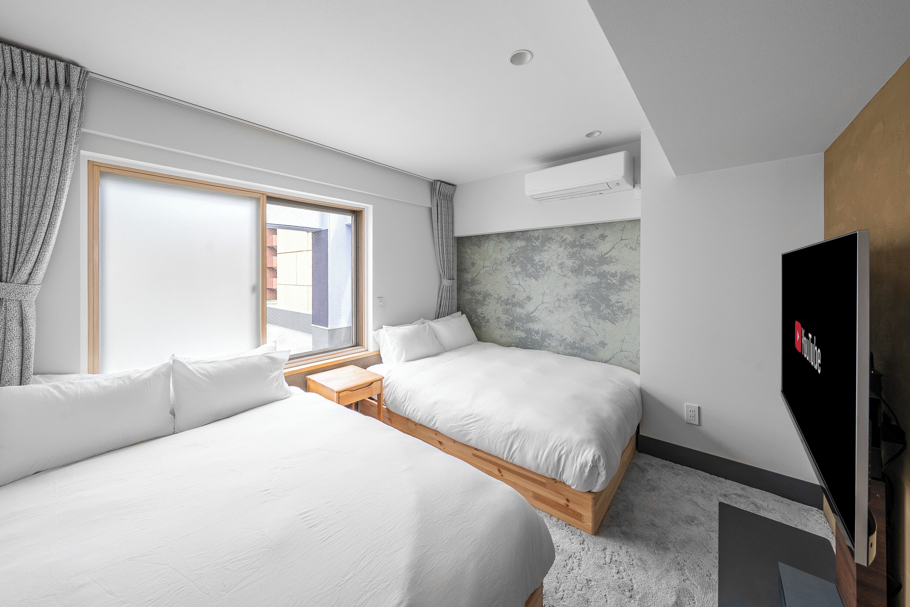 A modern hotel room with two beds, a large window, an air conditioning unit, and a flat-screen TV.