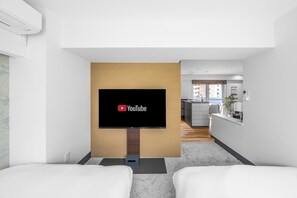 A modern hotel room with a flat-screen TV mounted on a wooden stand, a bed, a small seating area, and a view of the kitchen and balcony. - MIMO (Sapporo)