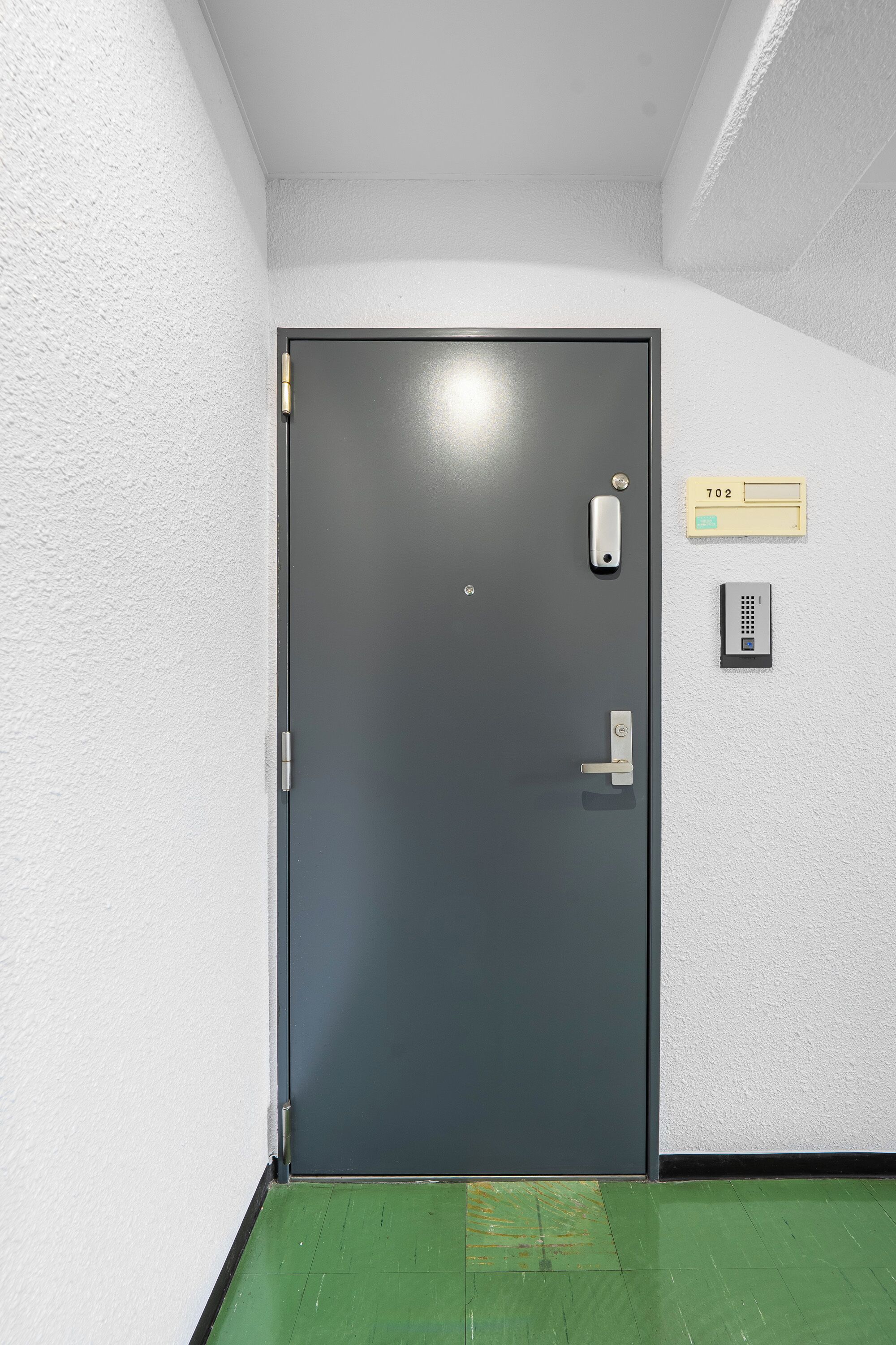 A closed door with a keypad lock, a doorbell, and a mailbox with the number 702 on a white wall.
