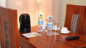 A wooden table with a black coffee maker, two water bottles, a glass, a white cup, and a remote control. - Blue Junction Eco Lodge (Arusha)