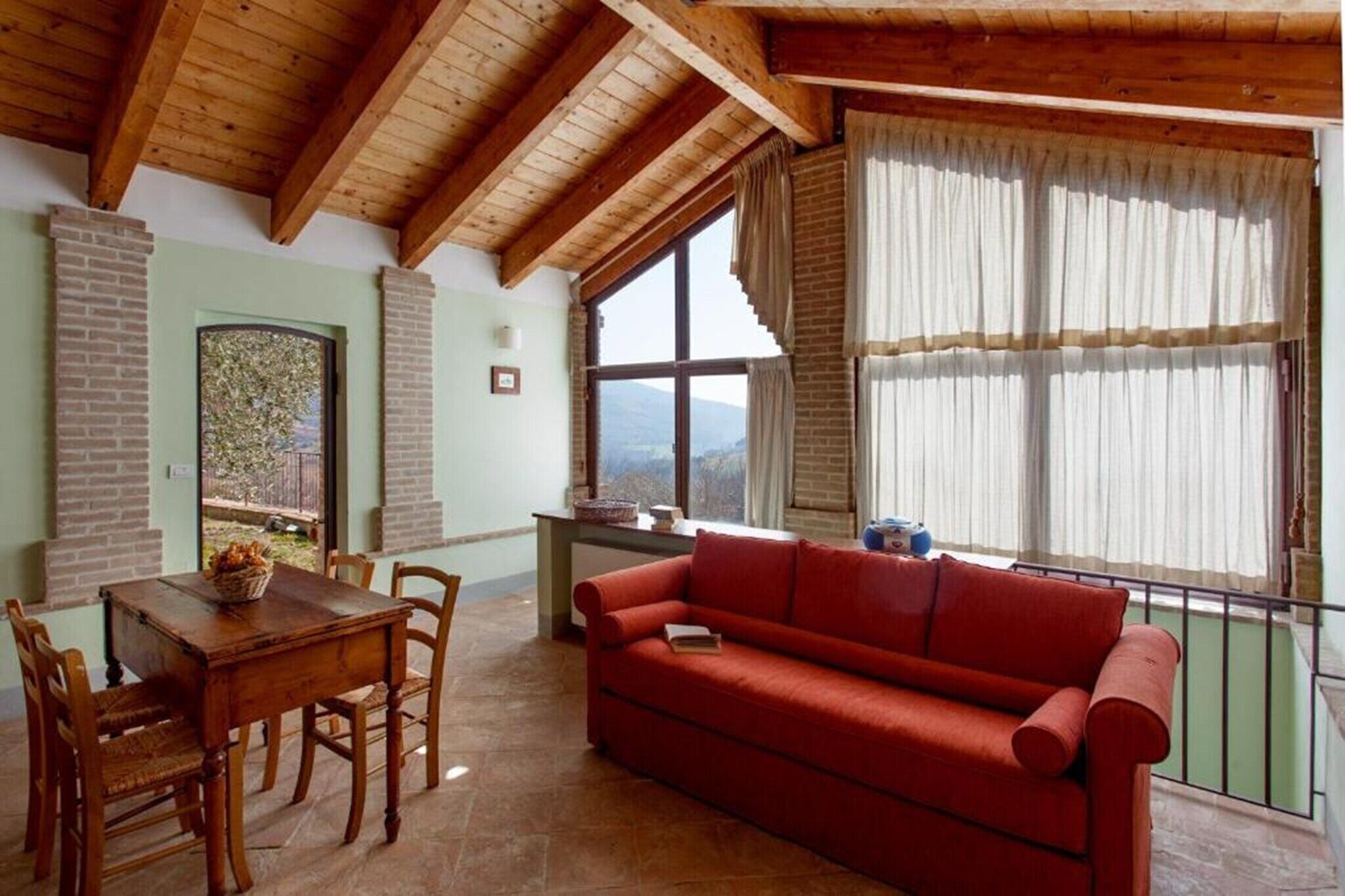 A cozy living room with a wooden ceiling, a red sofa, a dining table with chairs, and a view of the outdoors through large windows.