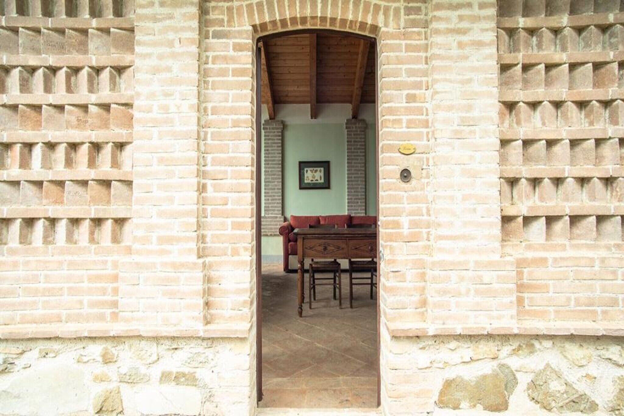 Un arc en briques mène à une pièce où se trouvent une table et des chaises, un tableau encadré au mur et un interphone.