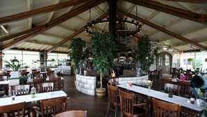 A spacious restaurant with high ceilings, wooden beams, and a central fire feature. Tables are set with chairs, and potted plants are placed around the room.