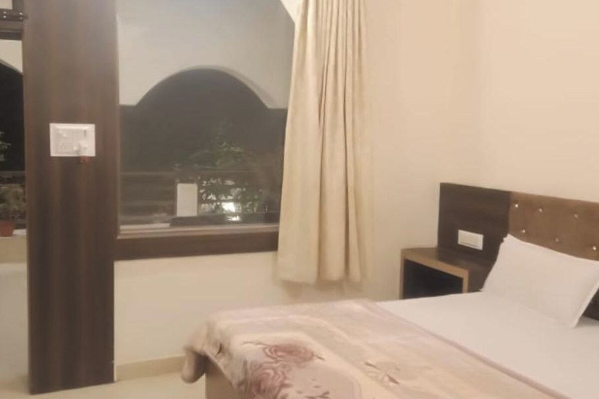 A hotel room with a bed, a nightstand, a window with curtains, and a view of the outdoors.