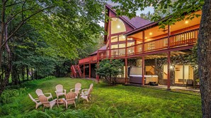 A two-story wooden cabin with a balcony, surrounded by lush greenery and a well-maintained lawn.