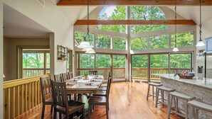 A spacious dining area with a long wooden table set for six, surrounded by wooden chairs. Large windows offer a view of the surrounding trees.