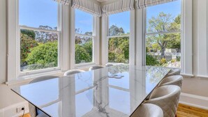 A dining area with a glass table and chairs, large windows with curtains, and a view of greenery outside. - Talina - History in the Heart of St Helens (Break O'Day)