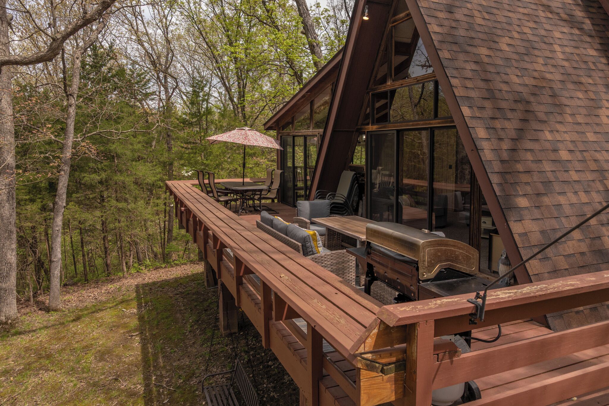 Une terrasse en bois avec des meubles d’extérieur, un barbecue et une grande fenêtre donnant sur une zone boisée.