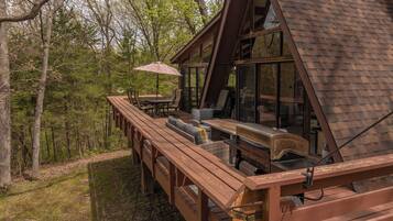 Une terrasse en bois avec des meubles d’extérieur, un barbecue et une grande fenêtre donnant sur une zone boisée.