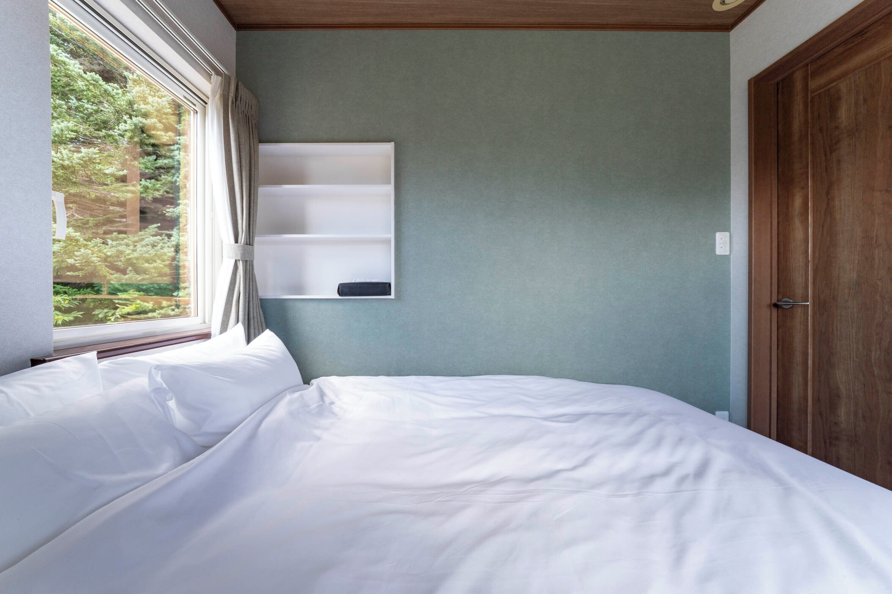 Une chambre à coucher avec un lit, une fenêtre avec des rideaux, une étagère fixée au mur et une porte en bois.