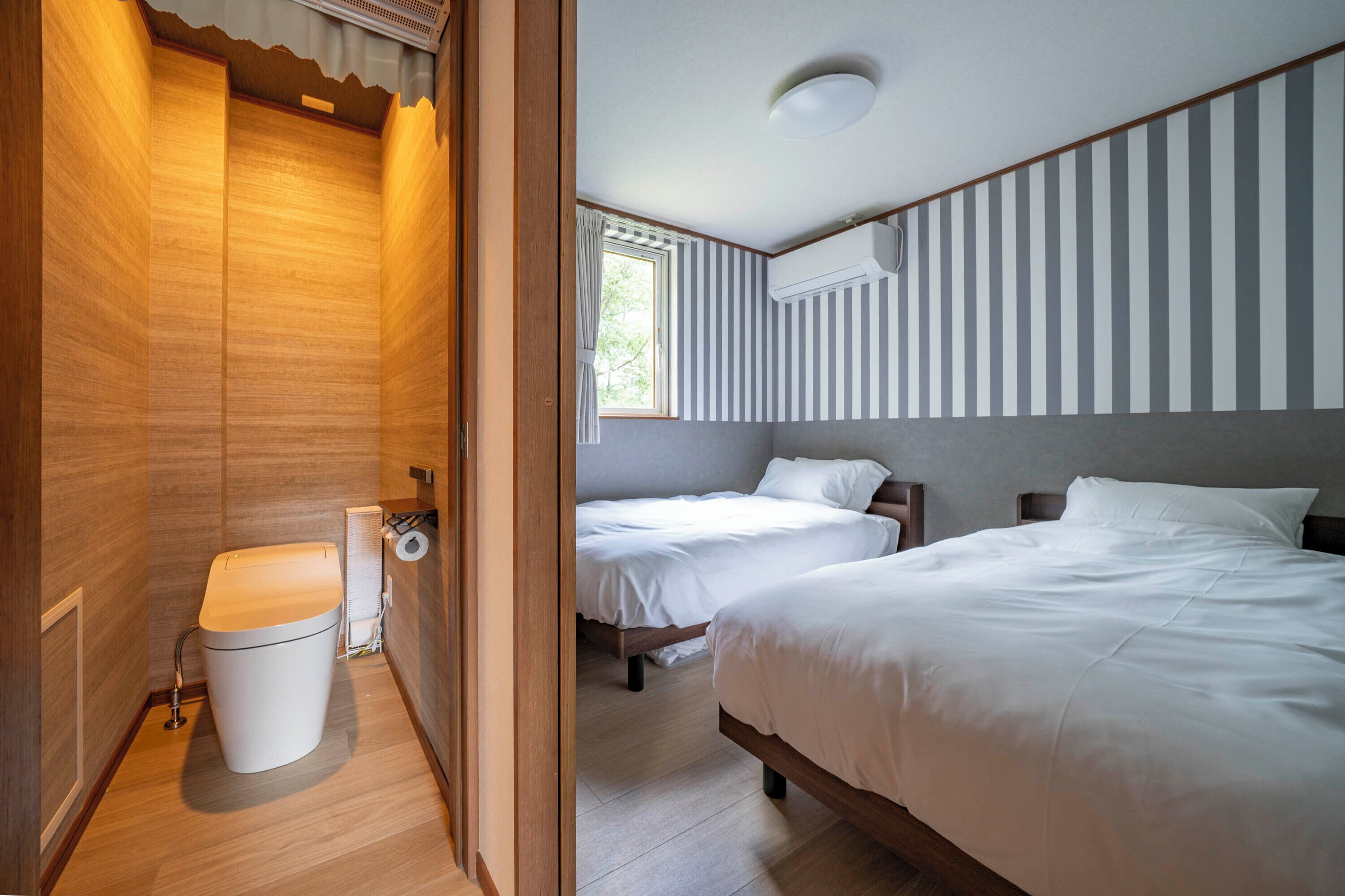 A hotel room with a wooden floor, two beds, a window with striped curtains, an air conditioning unit, and a toilet.