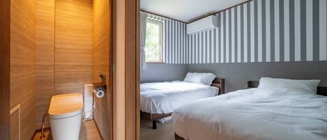 A hotel room with a wooden floor, two beds, a window with striped curtains, an air conditioning unit, and a toilet.