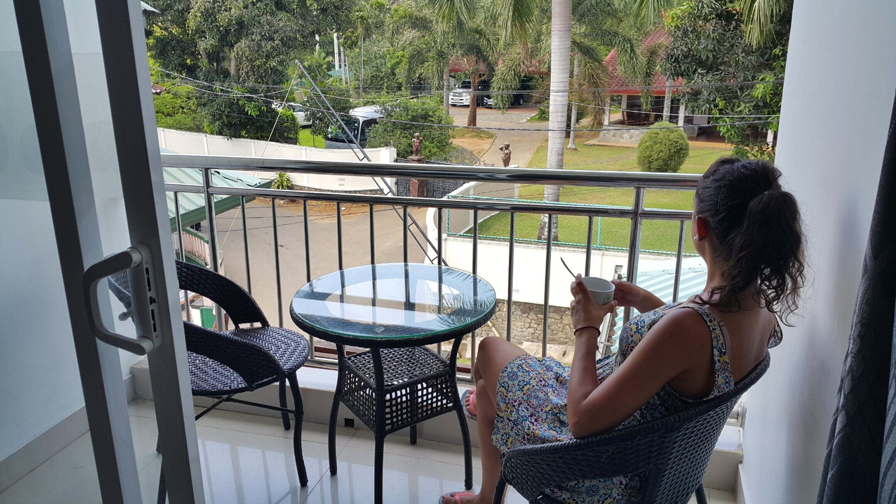 A balcony with a glass table and wicker chairs, overlooking a street with parked cars and greenery.