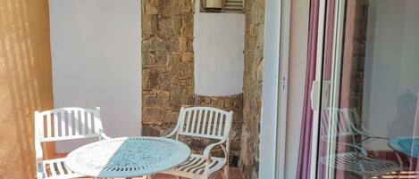 Un pequeño patio con una mesa redonda y cuatro sillas blancas, un muro de piedra y una ventana con cortinas.