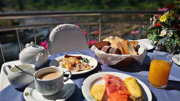 Una mesa dispuesta para el desayuno con tostadas, huevos, tocino y fruta, con vistas a una masa de agua al fondo.