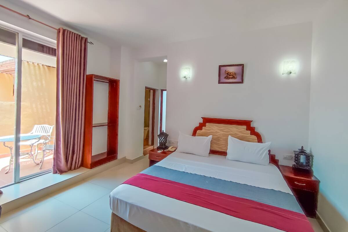 A hotel room with a bed, a nightstand, a wall-mounted lamp, and a sliding glass door leading to an outdoor area with chairs.